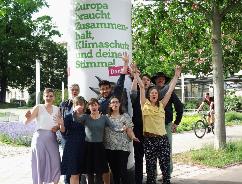 Grüne Stadtratskandidat*innen vor einer Litfaßsäule mit Europawahlmotiv und Störer "Danke".